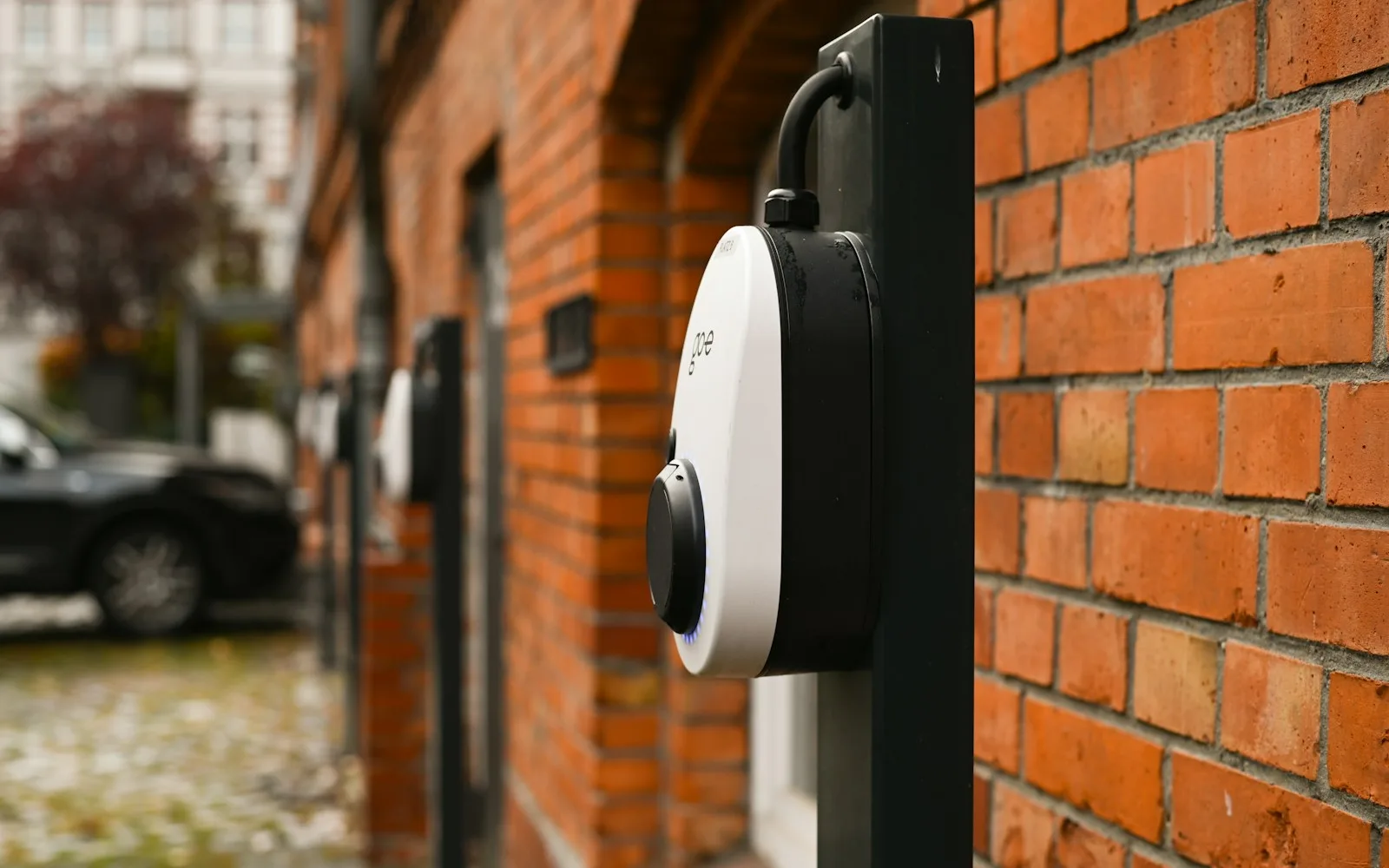 Fila di wallbox per la ricarica elettrica installate su colonnine lungo il parcheggio di un edificio residenziale in mattoni.